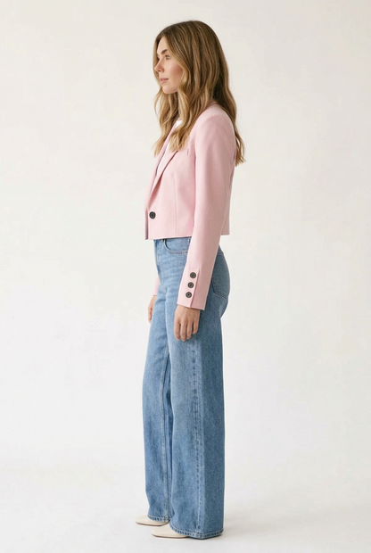 A woman modeling a light pink cropped blazer with black buttons, worn over a white t-shirt and blue jeans, against a plain off-white studio background.