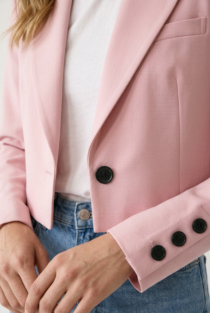 A woman modeling a light pink cropped blazer with black buttons, worn over a white t-shirt and blue jeans, against a plain off-white studio background.