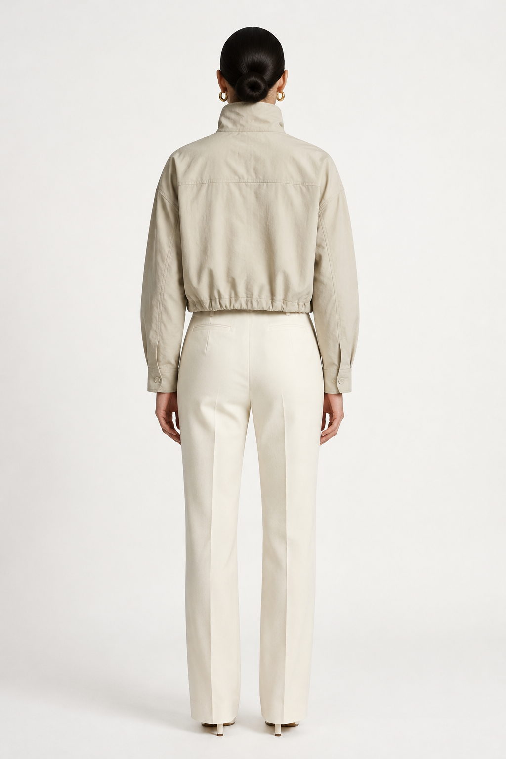 A model wearing a light beige cropped jacket with a high mock neck and drawstring details, paired with cream-colored trousers, against a plain off-white studio background.