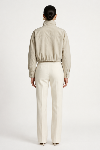 A model wearing a light beige cropped jacket with a high mock neck and drawstring details, paired with cream-colored trousers, against a plain off-white studio background.