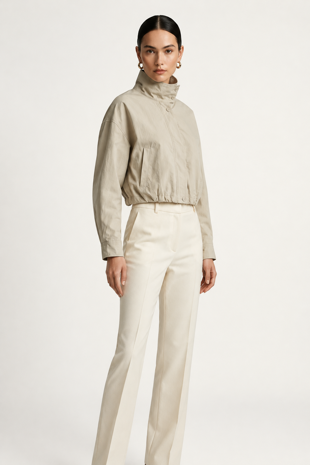 A model wearing a light beige cropped jacket with a high mock neck and drawstring details, paired with cream-colored trousers, against a plain off-white studio background.