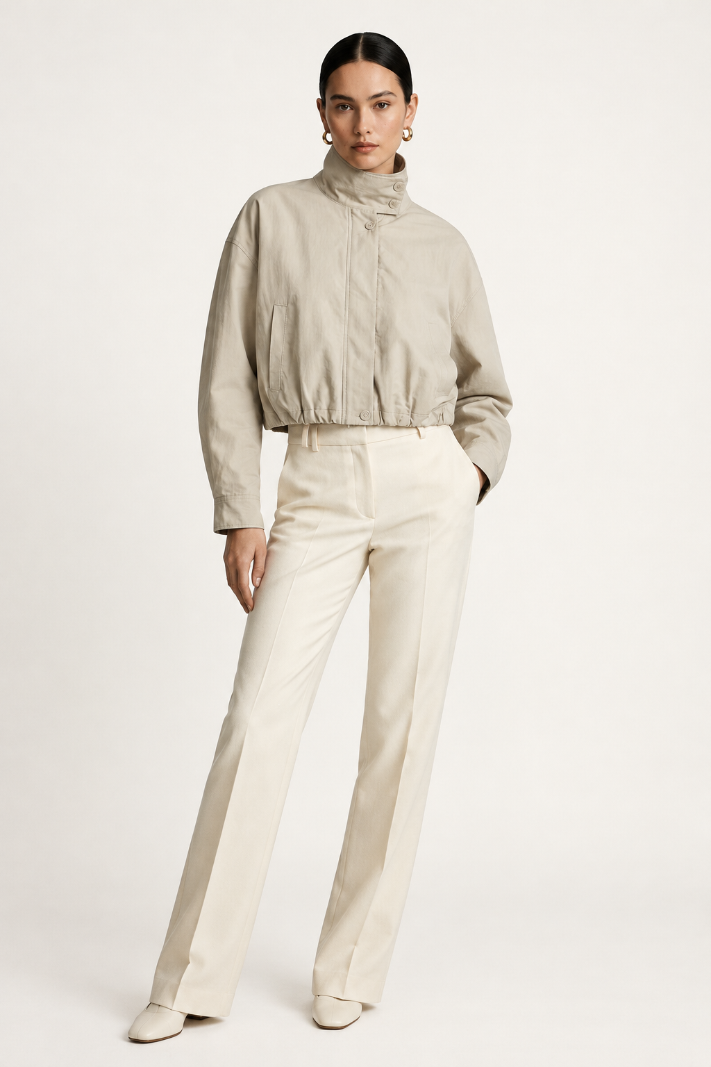 A model wearing a light beige cropped jacket with a high mock neck and drawstring details, paired with cream-colored trousers, against a plain off-white studio background.