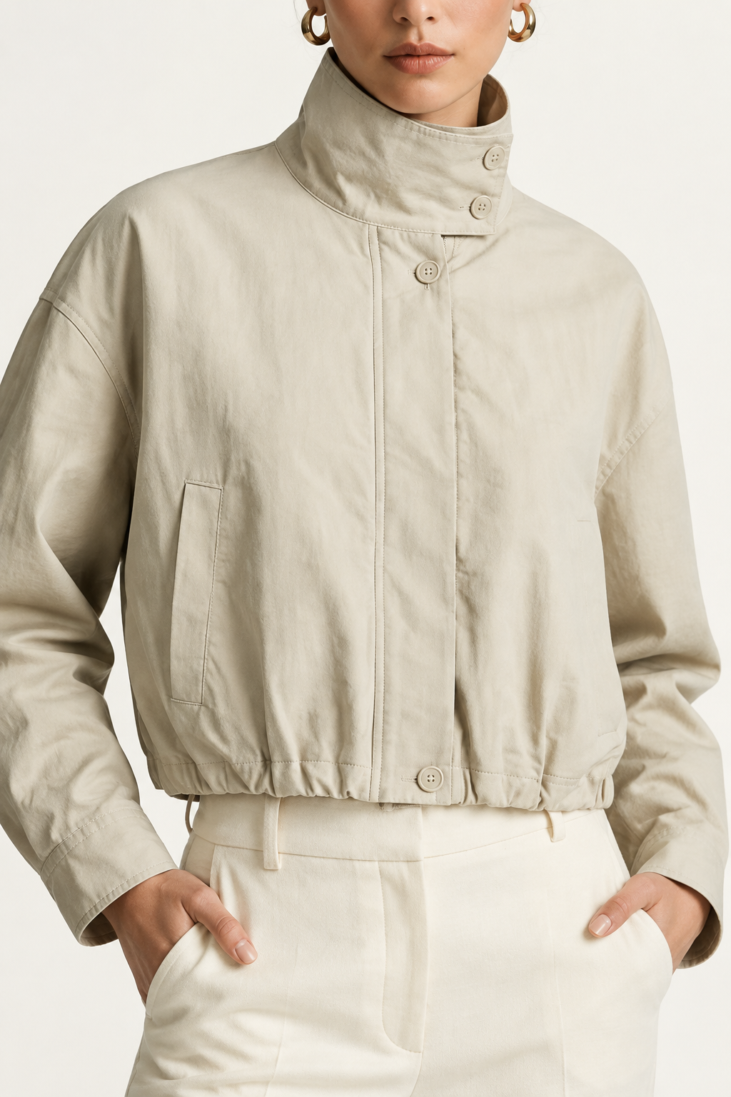 A model wearing a light beige cropped jacket with a high mock neck and drawstring details, paired with cream-colored trousers, against a plain off-white studio background.