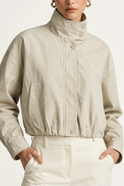 A model wearing a light beige cropped jacket with a high mock neck and drawstring details, paired with cream-colored trousers, against a plain off-white studio background.