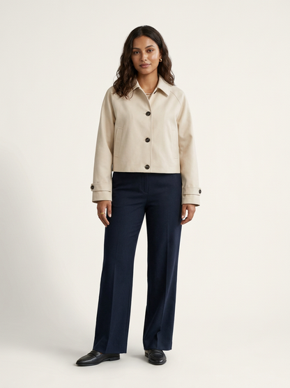 A woman modeling a cropped beige button-front jacket with dark buttons, paired with navy wide-leg trousers and black shoes, against a plain light beige studio background.