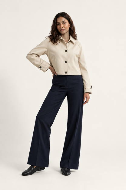 A woman modeling a cropped beige button-front jacket with dark buttons, paired with navy wide-leg trousers and black shoes, against a plain light beige studio background.