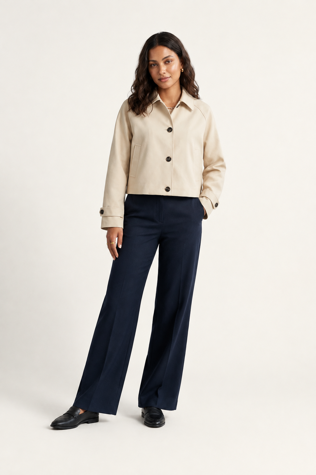 A woman modeling a cropped beige button-front jacket with dark buttons, paired with navy wide-leg trousers and black shoes, against a plain light beige studio background.