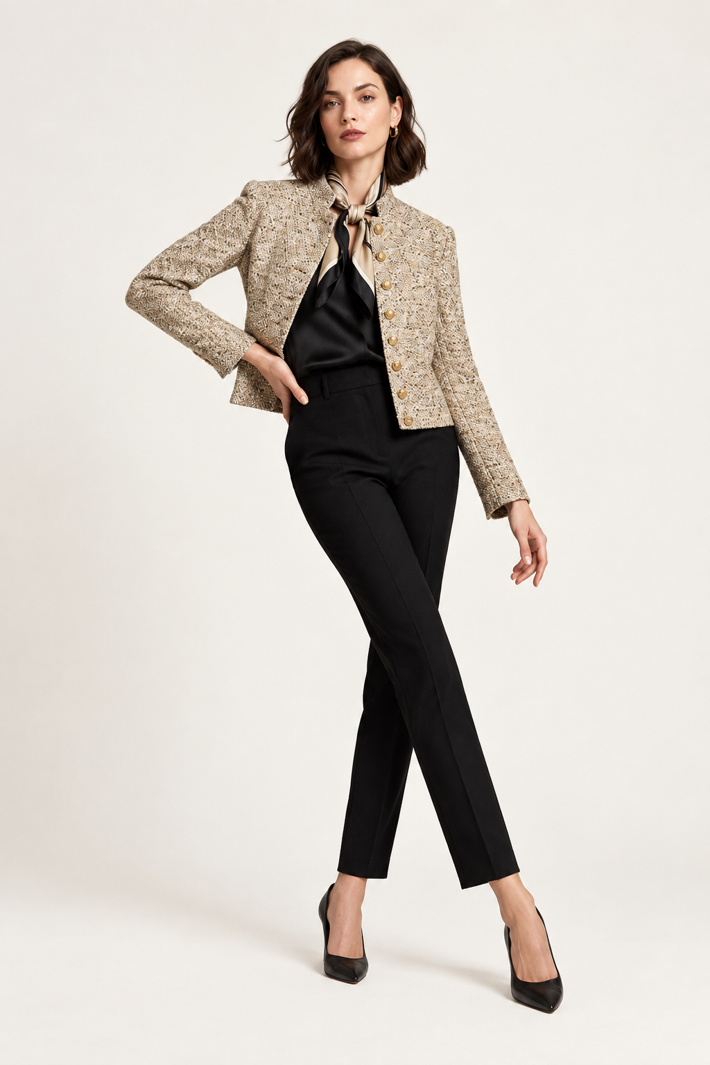 A model poses in a studio wearing a cropped beige tweed jacket with gold buttons, a black satin necktie blouse, black trousers, and black heels against a light gray background.