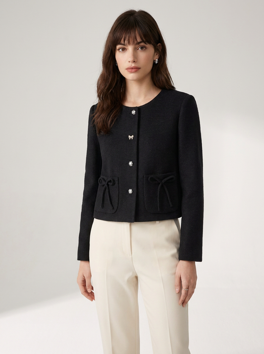 A woman modeling a black cropped tweed jacket with decorative silver buttons and drawstring details on the front pockets, paired with cream-colored trousers, against a neutral studio background.