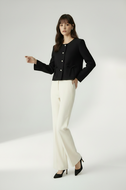 A woman modeling a black cropped jacket with silver-tone buttons and a tie detail at the waist, paired with off-white wide-leg trousers and black pointed heels, against a light gray studio background.