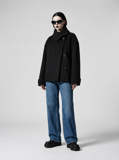 A model wearing a black double-breasted coat, blue wide-leg jeans, and black loafers, standing against a light gray studio background.