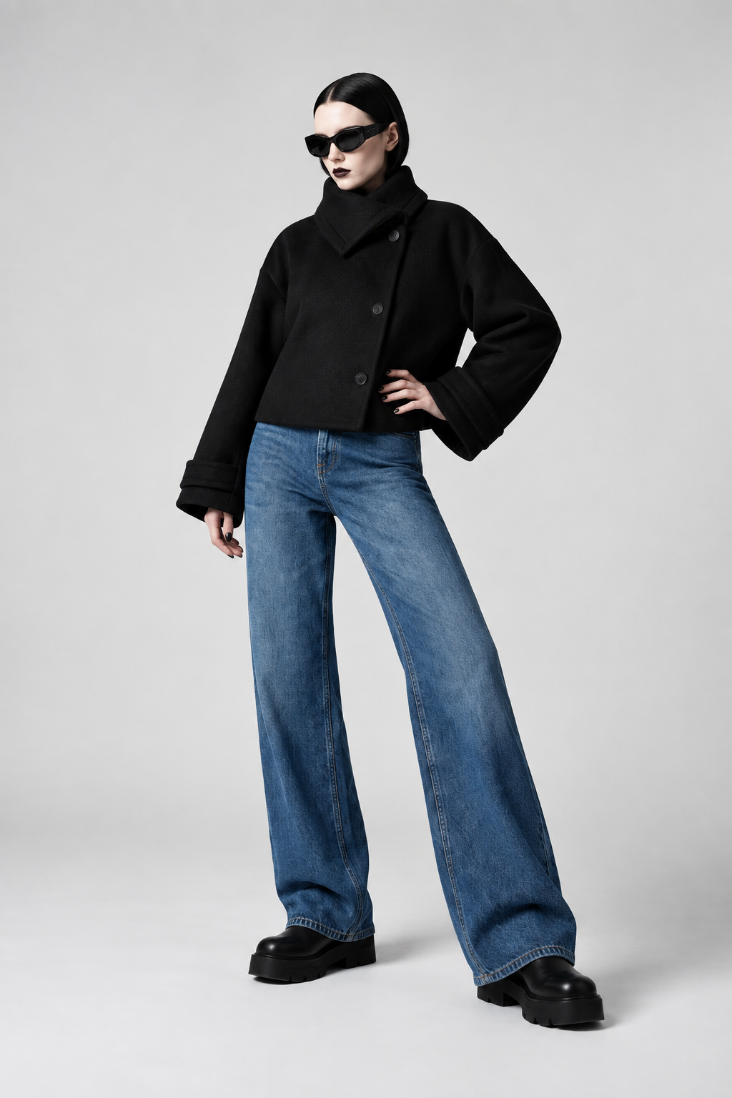 A model wearing a black cropped jacket, wide-leg blue jeans, and black platform shoes, posing against a light gray studio background.