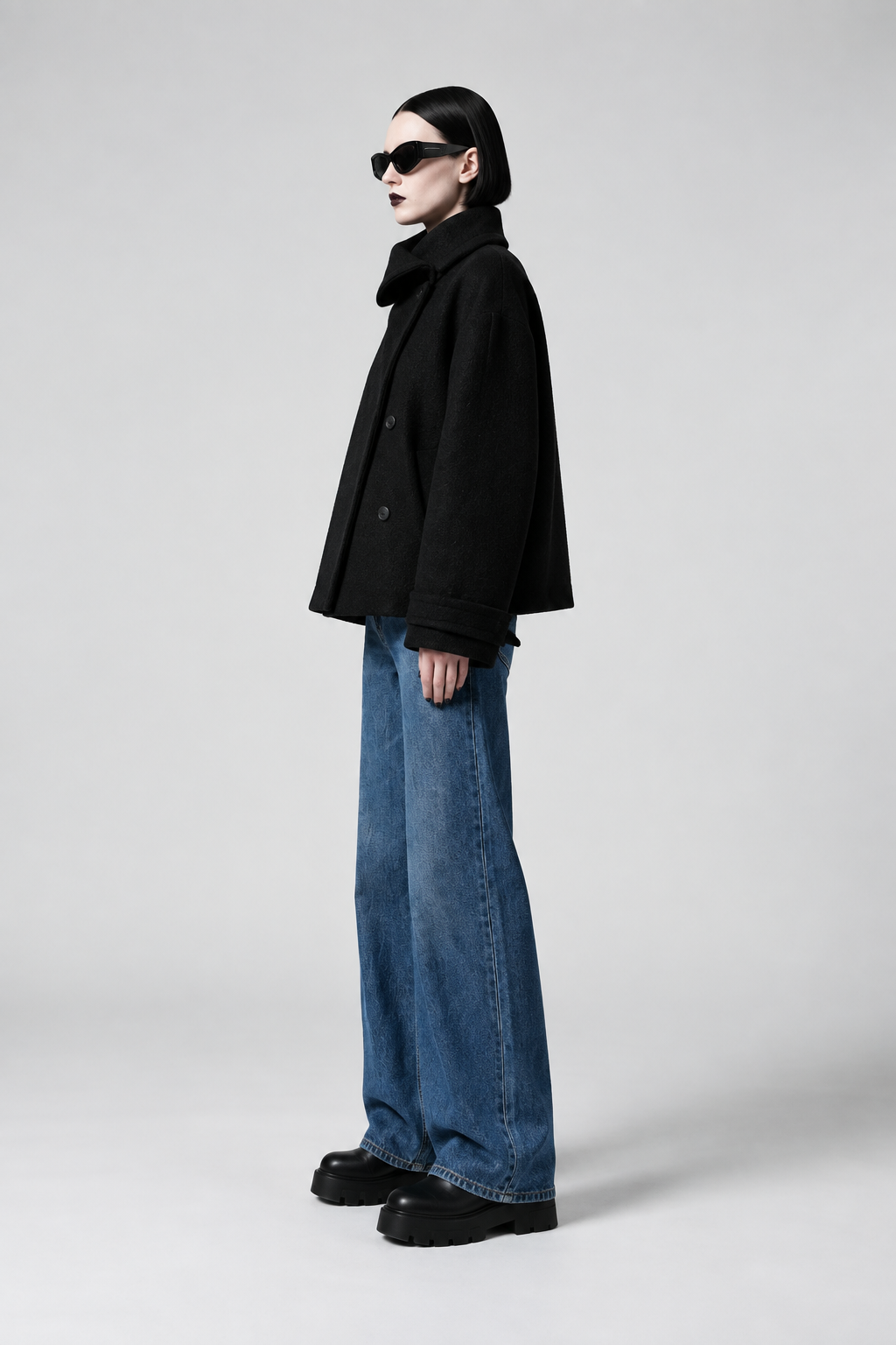 A model in profile wearing a black double-breasted coat, wide-leg blue jeans, black platform shoes, and black sunglasses against a light gray studio background.
