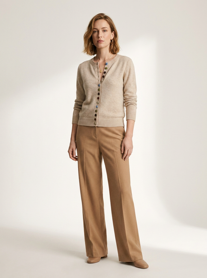 A woman stands in a studio wearing a light beige cardigan with decorative buttons and tan wide-leg trousers against a warm off-white background.