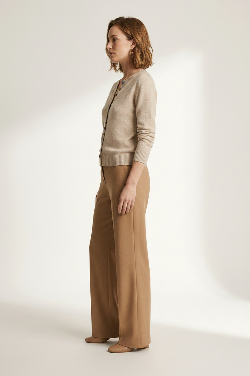 A woman stands in a studio wearing a light beige cardigan with decorative buttons and tan wide-leg trousers against a warm off-white background.