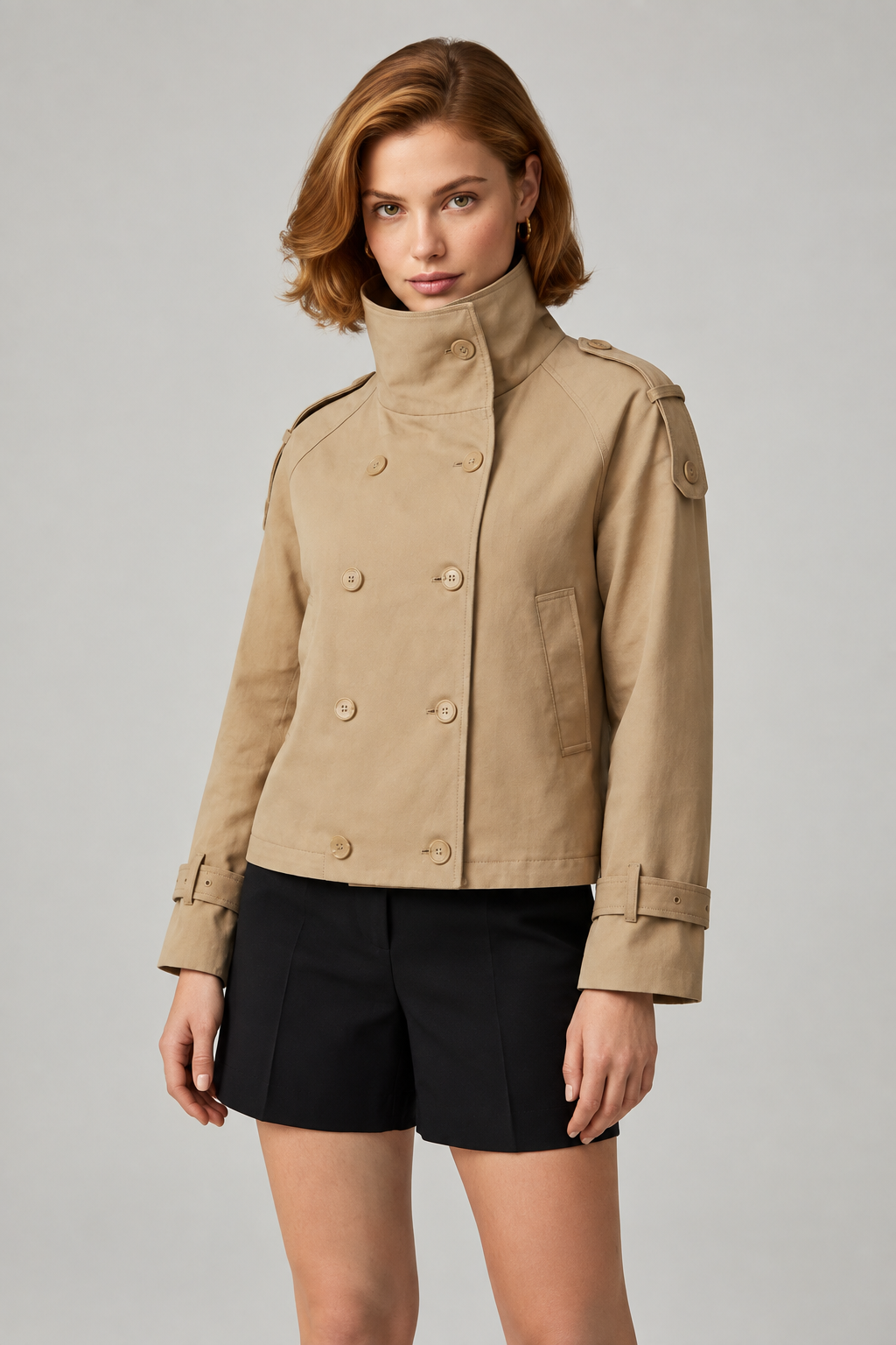 A woman modeling a cropped tan trench-style jacket with a high stand collar, double-breasted buttons, and shoulder epaulets, paired with black shorts, against a light gray studio background.