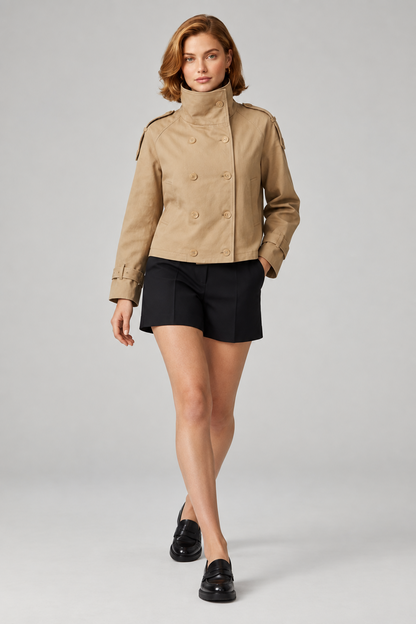 A model wearing a beige double-breasted cropped trench jacket with a high stand collar, paired with black shorts and black loafers, standing against a light gray studio background.