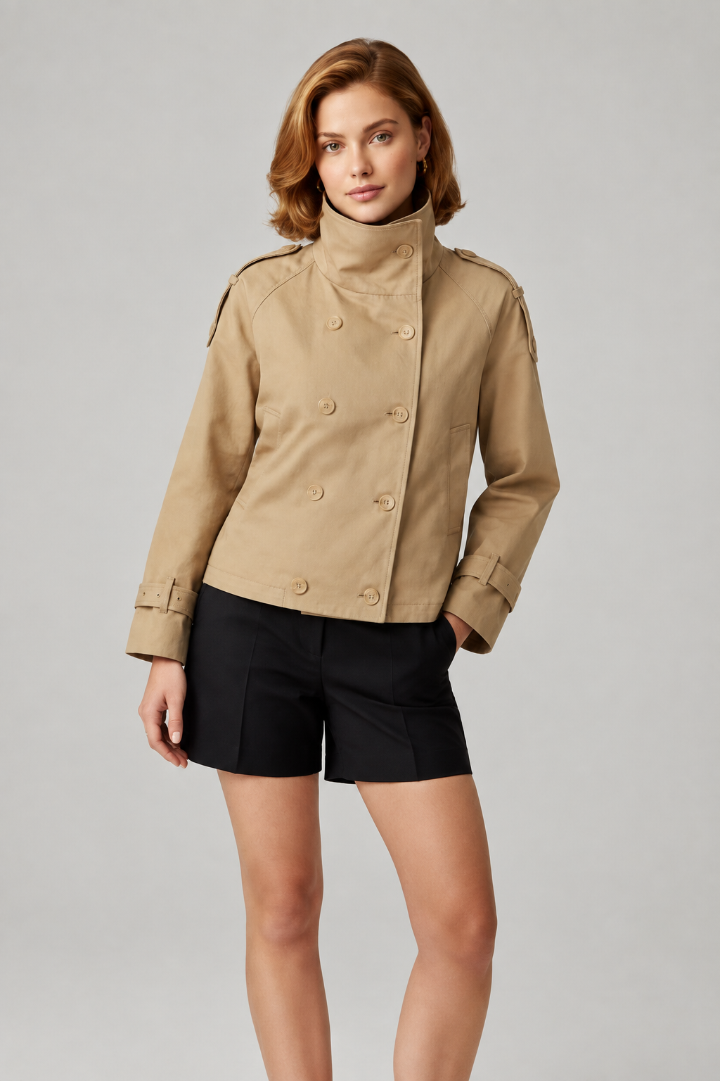 A woman modeling a tan double-breasted military-style jacket with a high collar and black shorts, photographed against a neutral light gray studio background.