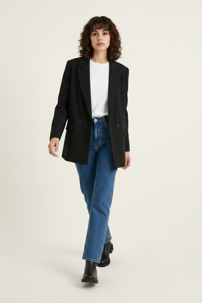 A woman modeling a black double-breasted blazer over a white crewneck top, paired with blue jeans and black ankle boots, against a light beige studio background.