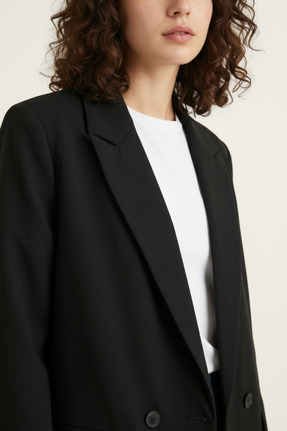 A close-up of a model wearing a black double-breasted blazer over a white crewneck top, set against a plain off-white background.