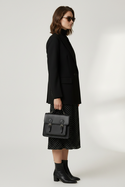 A model stands against a light gray studio background wearing a black double-breasted blazer over a black turtleneck, a black midi skirt with small white polka dots, and holding a black structured handbag.