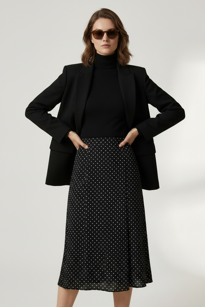 A model stands against a light gray studio background wearing a black double-breasted blazer over a black turtleneck, a black midi skirt with small white polka dots, and holding a black structured handbag.