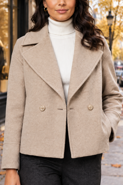 A woman wearing a beige double-breasted wool-blend coat over a white ribbed turtleneck sweater and dark gray trousers, photographed outdoors on a city street with blurred autumn trees in the background.