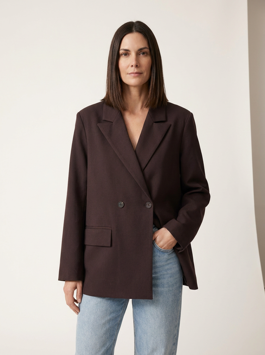 A woman modeling an oversized dark brown double-breasted blazer with light-wash blue jeans, standing against a plain off-white studio background.