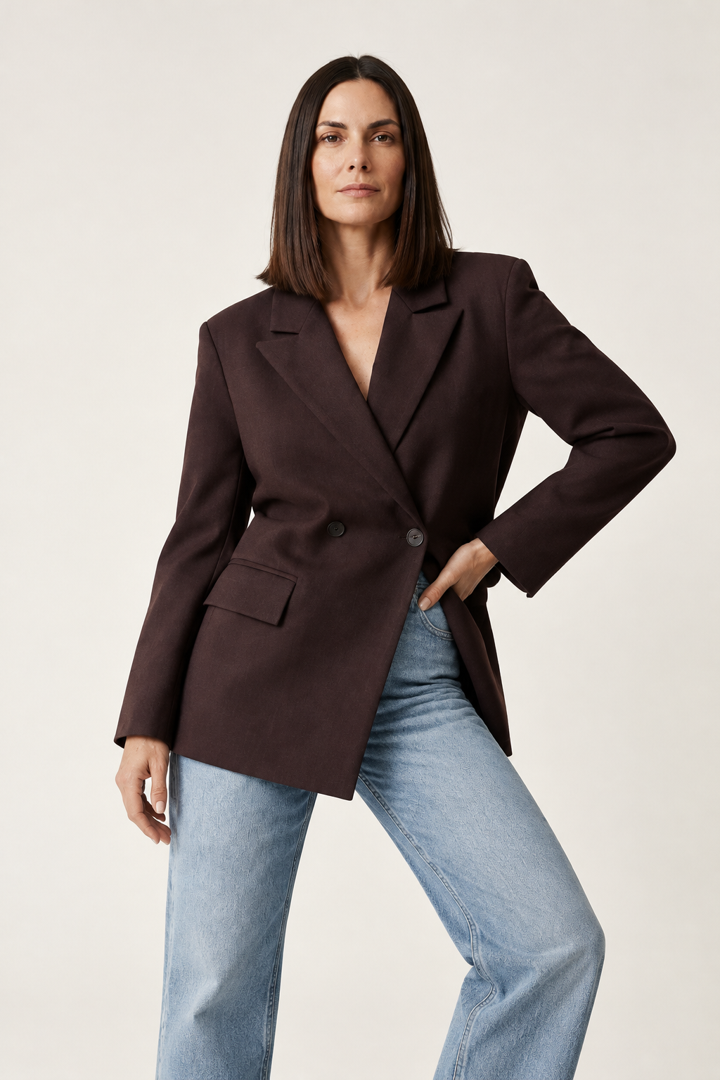 A woman modeling an oversized dark brown double-breasted blazer with light-wash blue jeans, standing against a plain off-white studio background.