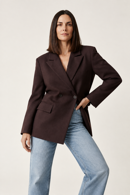 A woman modeling an oversized dark brown double-breasted blazer with light-wash blue jeans, standing against a plain off-white studio background.