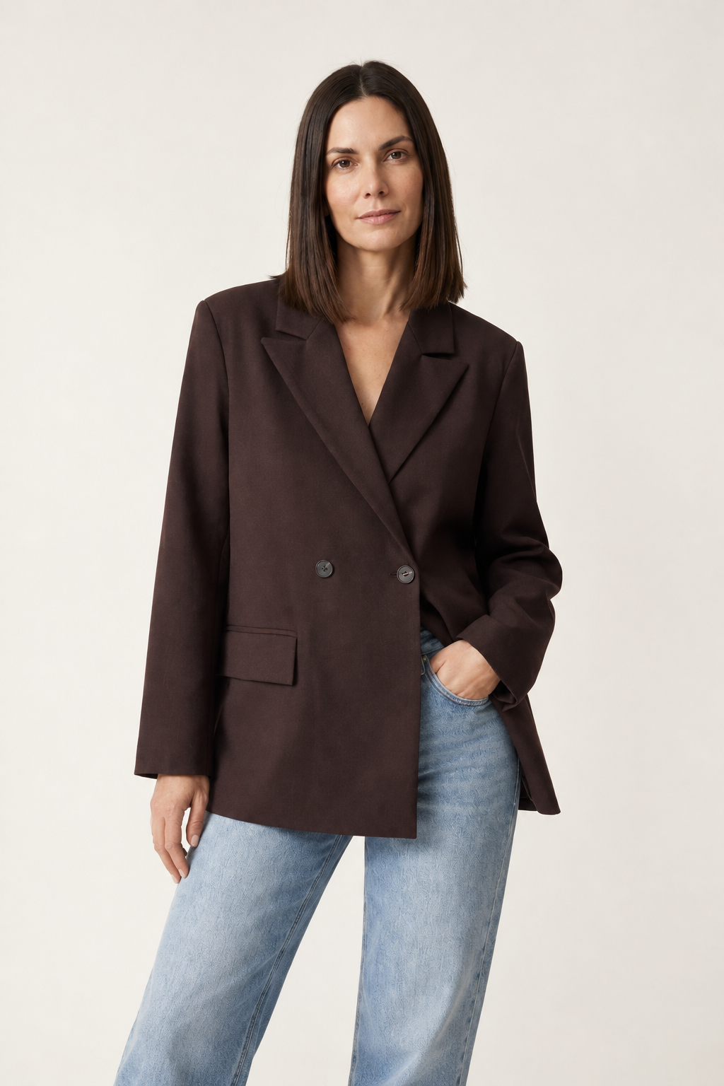 A woman modeling an oversized dark brown double-breasted blazer with light-wash blue jeans, standing against a plain off-white studio background.
