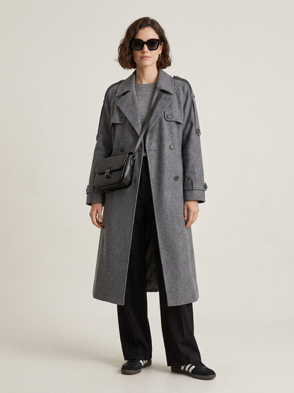A model wearing a long gray double-breasted coat over a gray top and black wide-leg trousers, accessorized with a black crossbody bag and black sneakers, photographed against a light gray studio background.