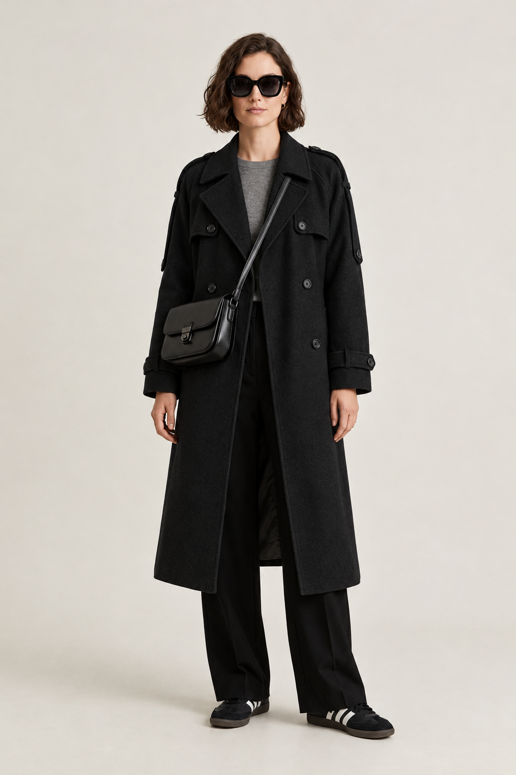 A model wearing a long gray double-breasted coat over a gray top and black wide-leg trousers, accessorized with a black crossbody bag and black sneakers, photographed against a light gray studio background.