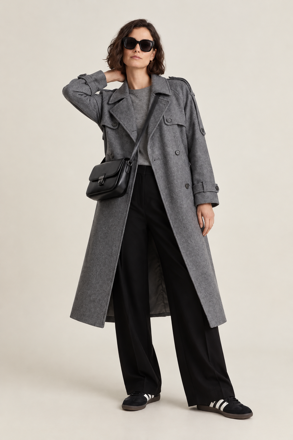 A model wearing a long gray double-breasted coat over a gray top and black wide-leg trousers, accessorized with a black crossbody bag and black sneakers, photographed against a light gray studio background.