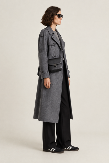 A model wearing a long gray double-breasted coat over a gray top and black wide-leg trousers, accessorized with a black crossbody bag and black sneakers, photographed against a light gray studio background.
