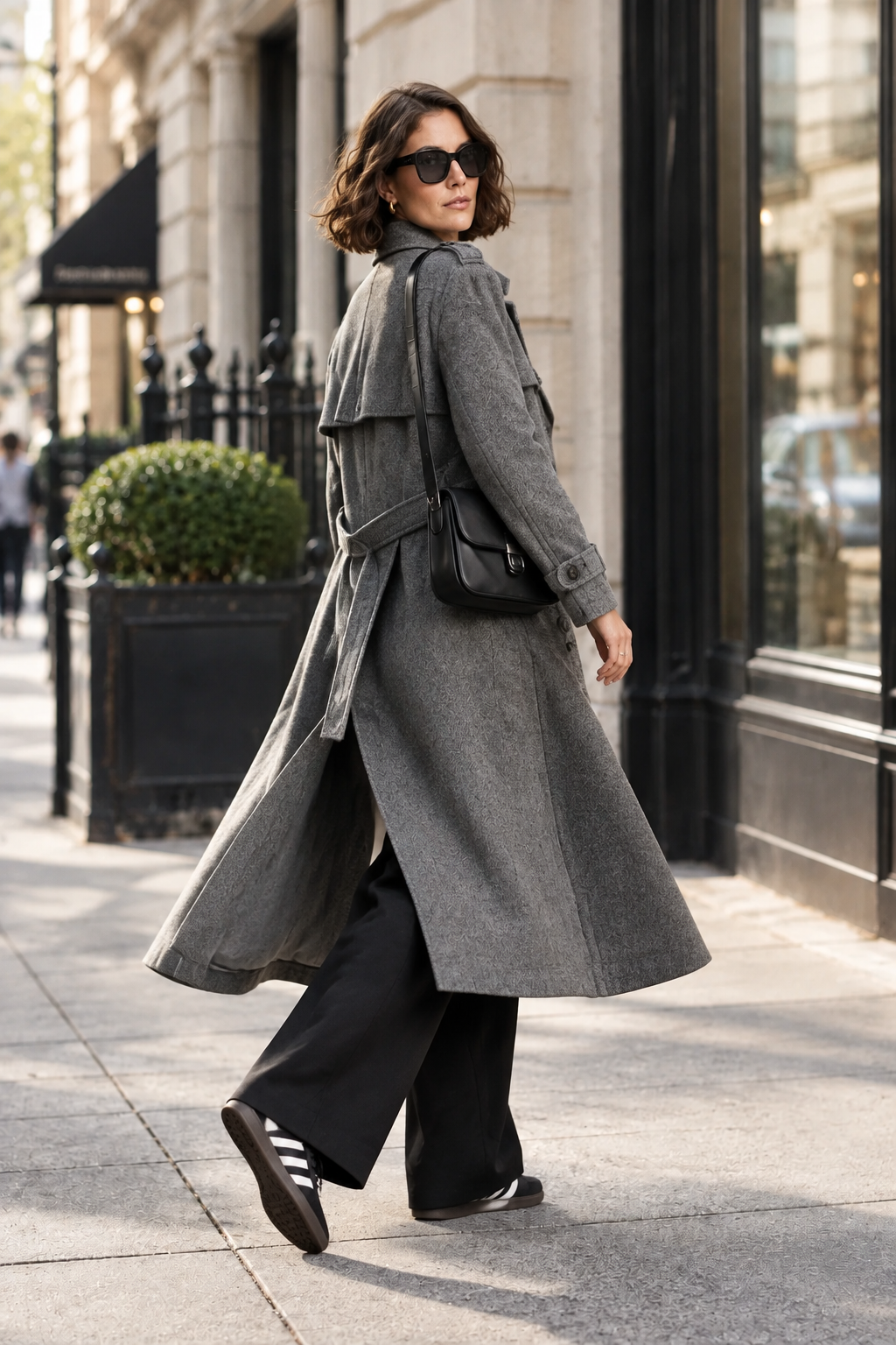 A woman stands on a city sidewalk wearing a long gray trench coat over a gray top, black wide-leg pants, black sneakers, and a black crossbody bag.