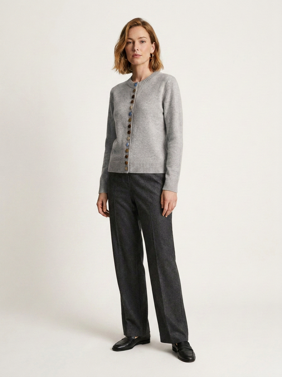 A woman modeling a light gray knit cardigan with dark buttons, paired with dark charcoal gray trousers and black shoes, against a plain off-white studio background.