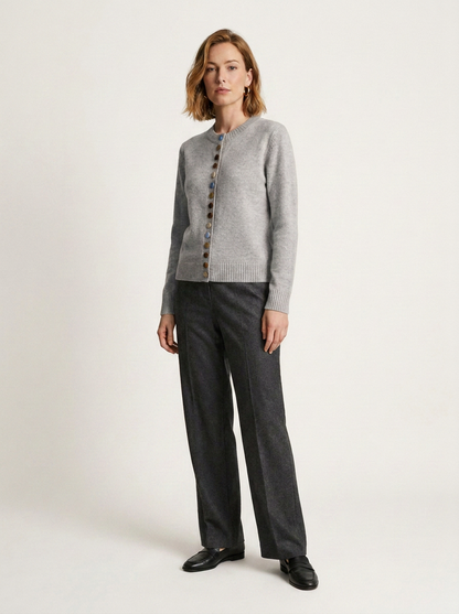 A woman modeling a light gray knit cardigan with dark buttons, paired with dark charcoal gray trousers and black shoes, against a plain off-white studio background.