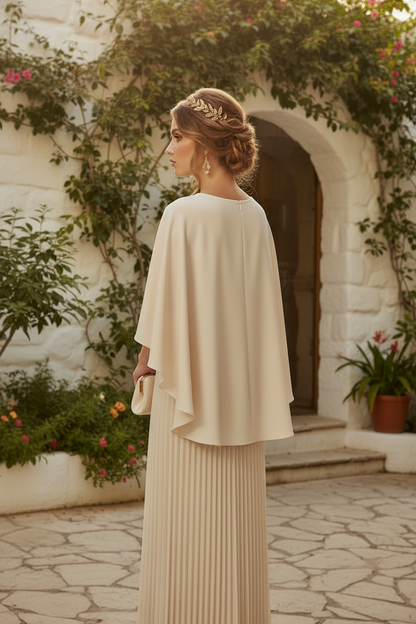 A woman stands outdoors in a sunlit courtyard wearing a cream-colored formal dress with a draped cape-style overlay and a pleated maxi skirt, holding a matching cream clutch.