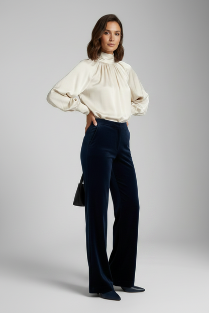A woman models a high-neck cream satin blouse and dark navy wide-leg velvet trousers, standing in profile against a neutral gray studio background.