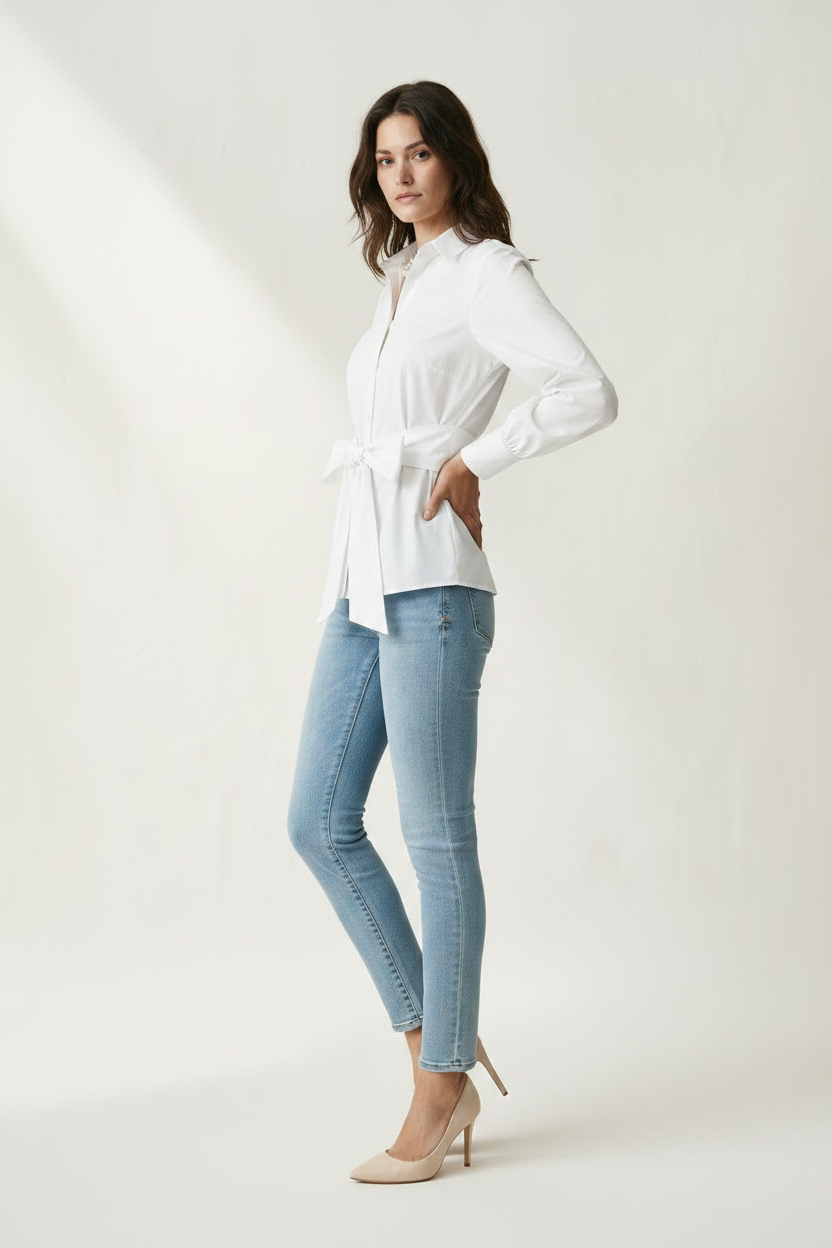A woman modeling a white long-sleeve button-up shirt with a tie waist, paired with light blue skinny jeans and beige heels, against a warm off-white studio background.