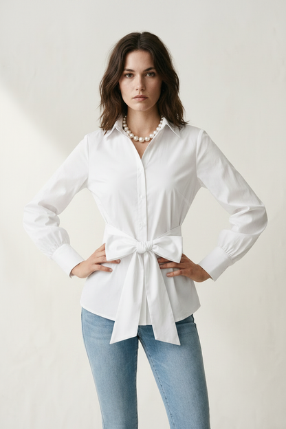 A woman modeling a white long-sleeve button-up shirt with a tie waist, paired with light blue jeans and a pearl necklace, against a warm off-white textured background.