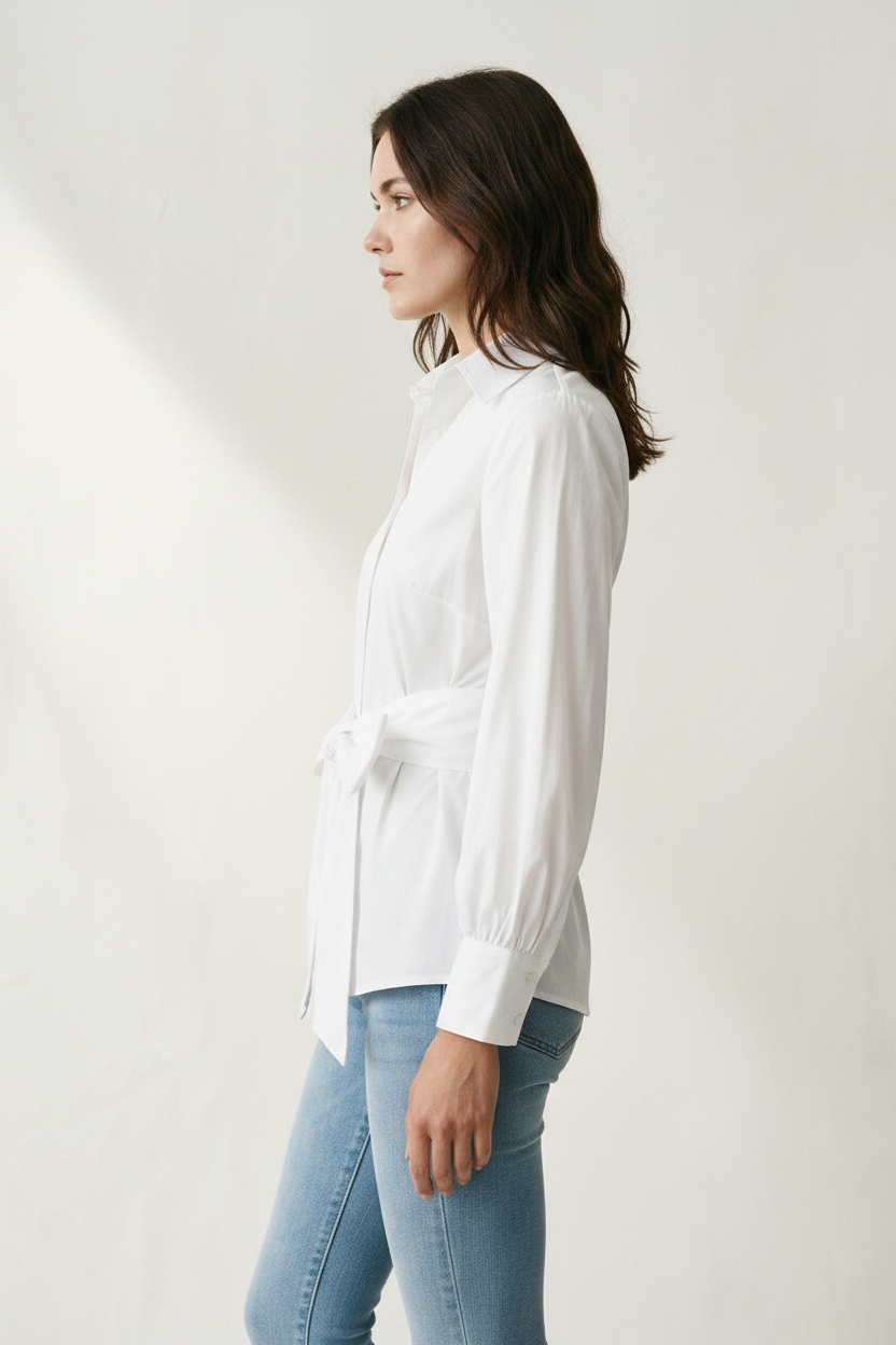A woman modeling a white long-sleeve button-up shirt with a tie waist, paired with light blue jeans and a pearl necklace, against a warm off-white textured background.