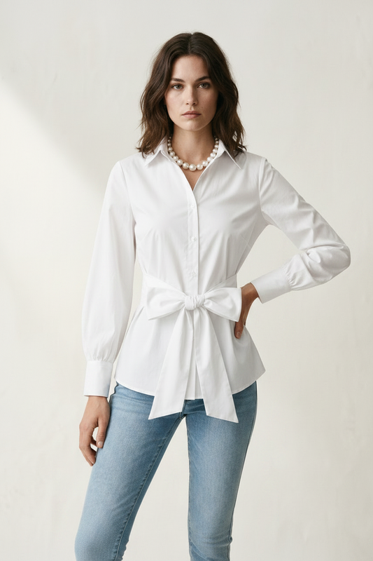 A woman modeling a white long-sleeve button-up shirt with a tie waist, paired with light blue jeans and a pearl necklace, against a warm off-white textured background.