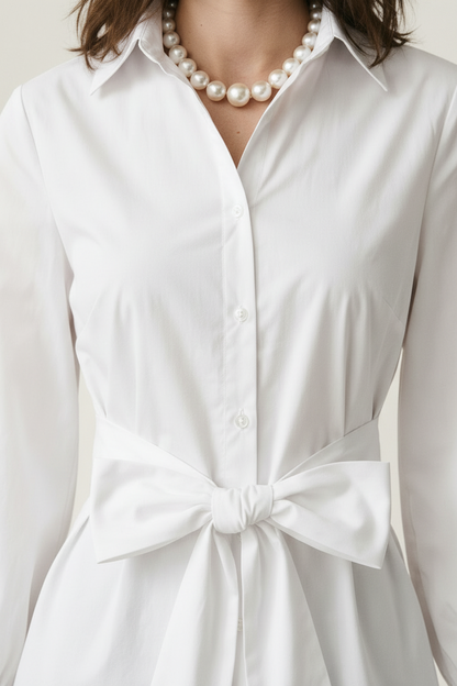 A woman modeling a white long-sleeve button-up shirt with a tie waist, paired with light blue jeans and a pearl necklace, against a warm off-white textured background.