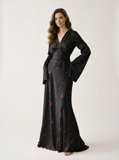 A woman modeling a long black satin-like maxi dress with a red floral print, featuring a wrap-style V-neckline, long flared sleeves, and a matching waistband, photographed against a plain light studio background.