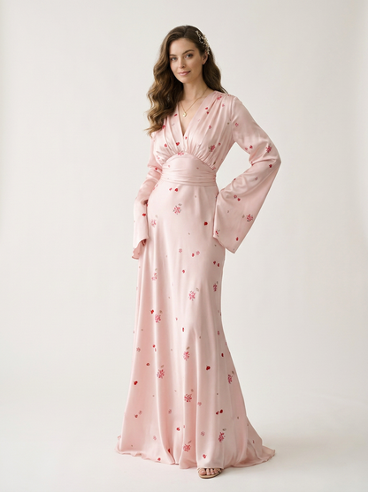 A woman modeling a long-sleeve, floor-length light pink gown with a subtle red floral pattern, photographed against a plain light gray studio background.