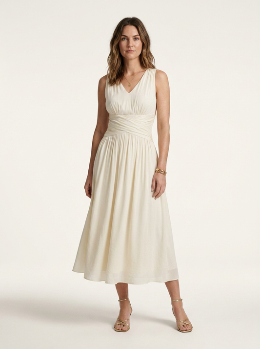 A woman modeling a sleeveless cream midi dress with a gathered waist and V-neckline, paired with gold strappy heels, against a plain light beige studio background.