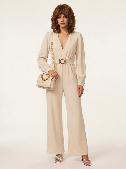 A woman models a beige long-sleeve wide-leg jumpsuit with a matching belt and gold-tone buckle, styled with a matching handbag and strappy heels against a plain light background.