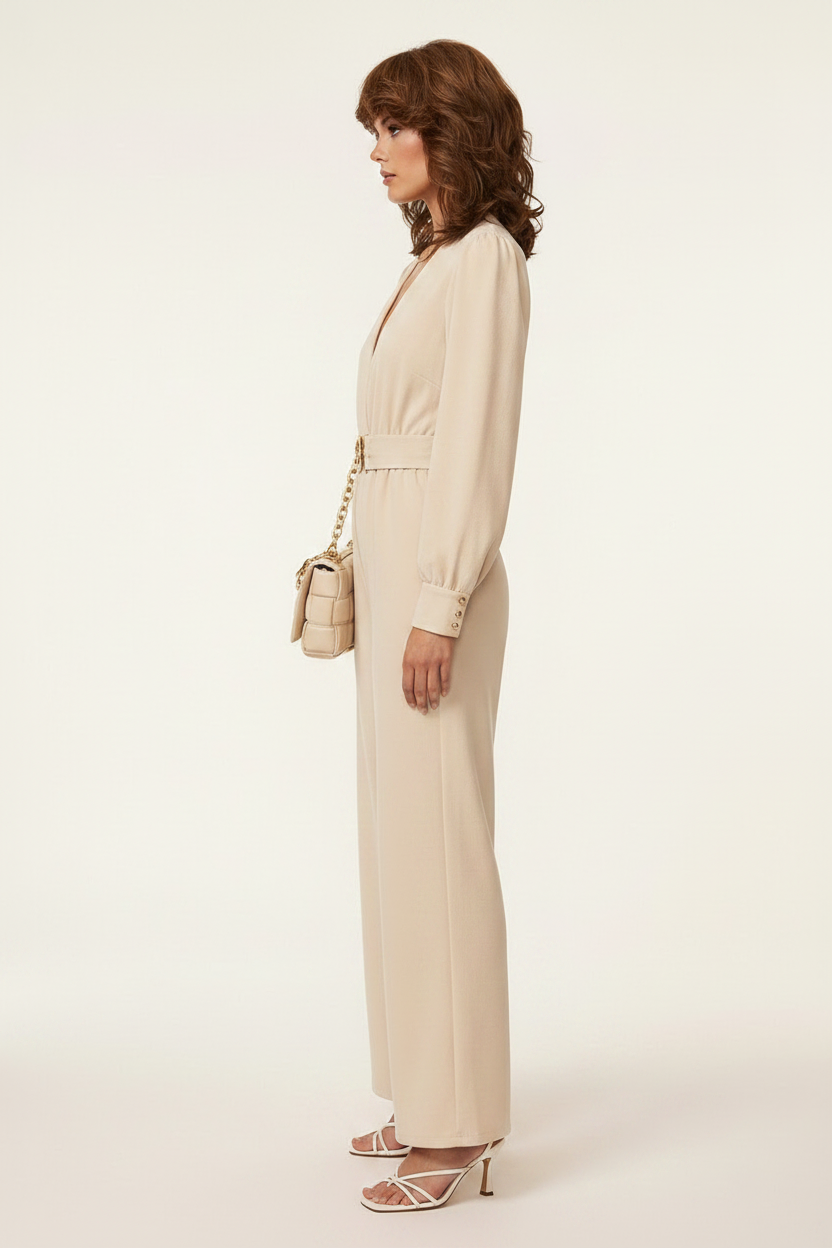A woman models a beige long-sleeve wide-leg jumpsuit with a matching belt and gold-tone buckle, styled with a matching handbag and strappy heels against a plain light background.